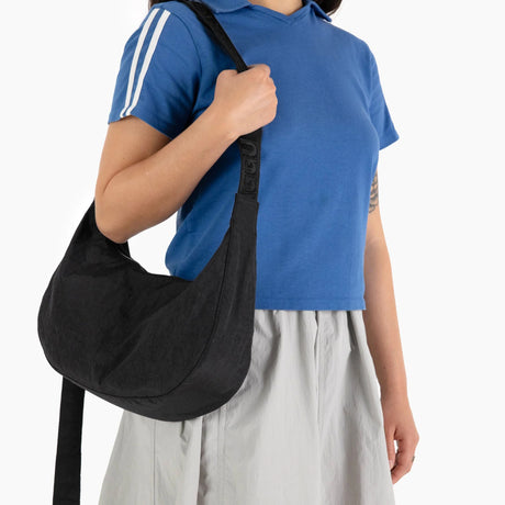 A person dressed in a blue shirt and gray skirt, carrying the Baggu Medium Nylon Crescent Bag in black with an adjustable strap, achieving a minimalist look.