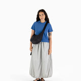 A person in a blue shirt and gray skirt stands with the Baggu Medium Nylon Crescent Bag - Black, showcasing its sleek design and adjustable strap for a minimalist look against the white background.