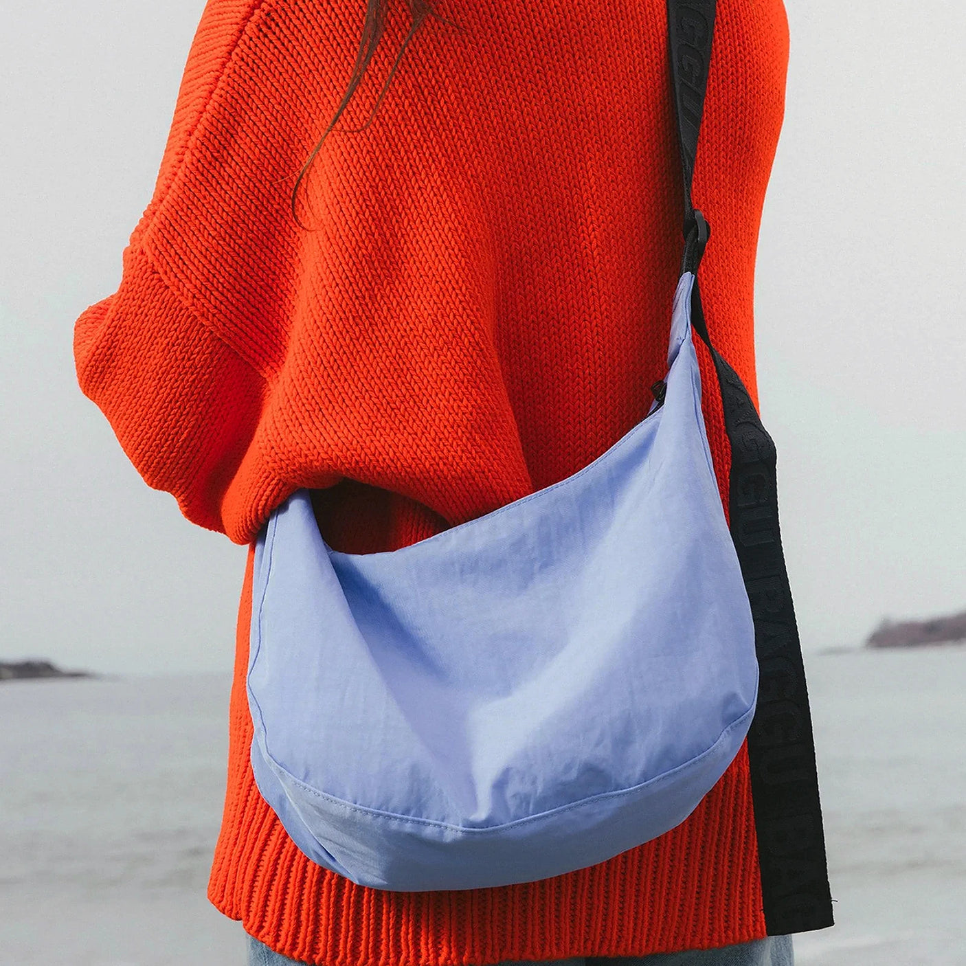 A person wearing an orange sweater with a Baggu Medium Nylon Crescent Bag in Cornflower, crafted from recycled nylon, stands by the waterfront.