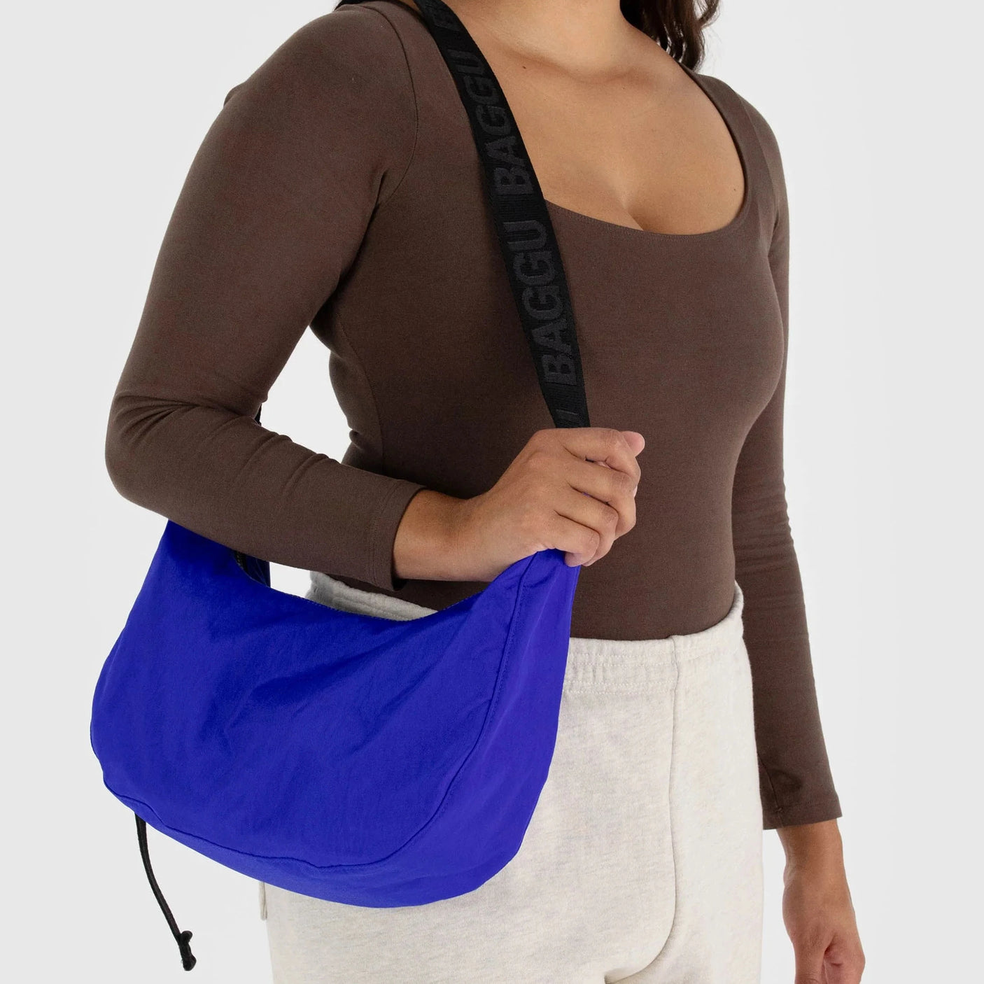 A person dressed in a brown top and white pants carries the Baggu Medium Nylon Crescent Bag in Lapis Blue, accented with a black strap. Crafted from recycled nylon, this bag introduces a vibrant touch to their outfit.