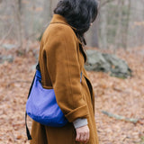 A person wearing a brown coat strolls through a forest carpeted with fallen leaves, carrying a Baggu Medium Nylon Crescent Bag in Lapis Blue, designed from recycled nylon.