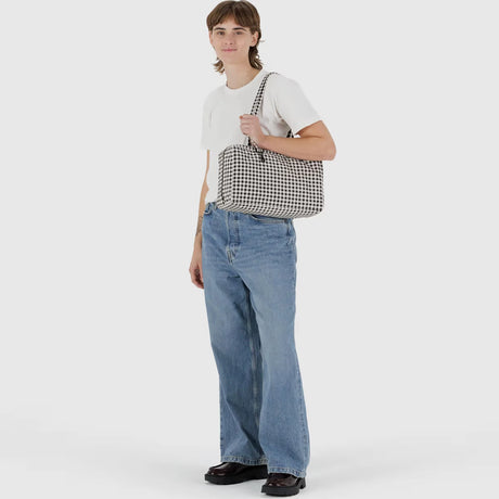 A person holds the Baggu Medium Nylon Bowler Bag - Gingham by Baggu, styled with a white t-shirt, loose blue jeans, and black shoes, against a plain light background.