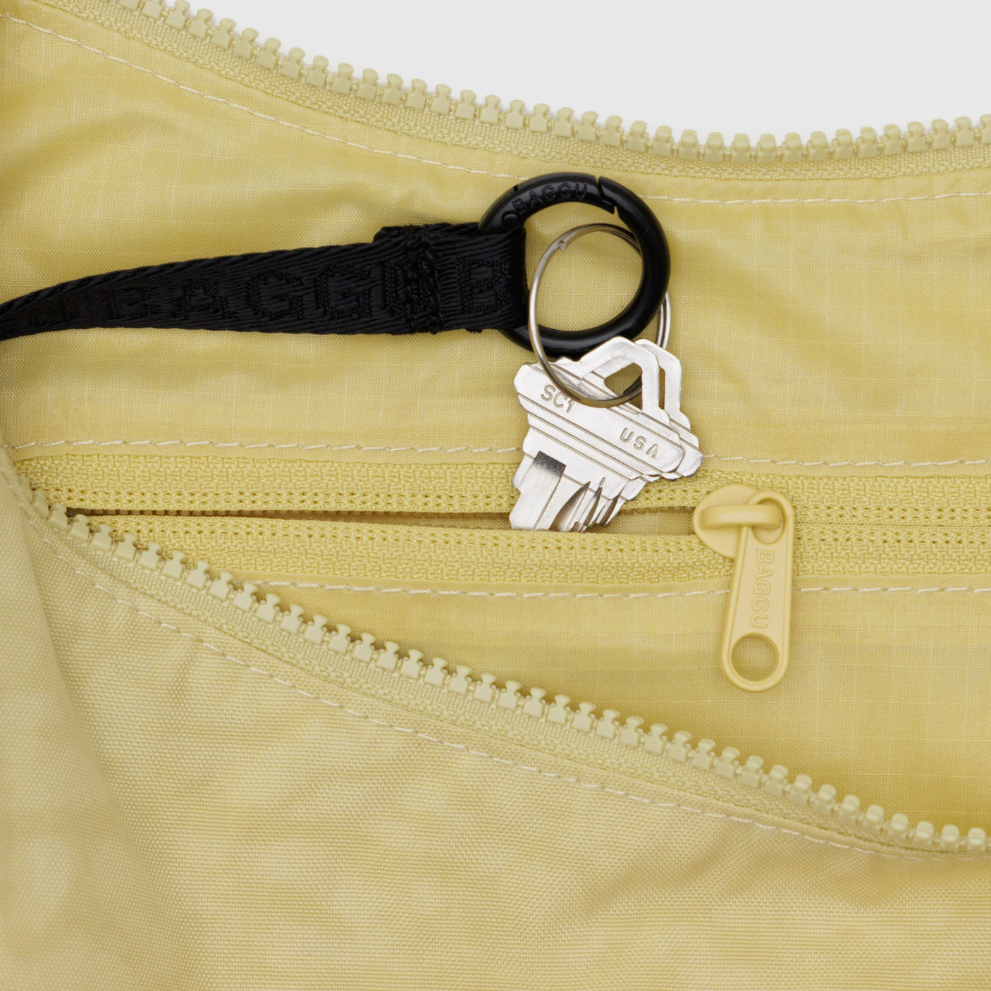 Keys attached to a black lanyard rest inside the open zippered pocket of a Baggu Medium Nylon Crescent Bag in Butter, which is machine washable.