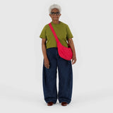 An older adult stands wearing a green shirt, wide blue jeans, and a Baggu Medium Nylon Crescent Bag in Candy Apple against a plain white background.