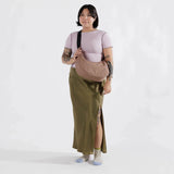 A tattooed person stands against a plain background, wearing a short-sleeved top, olive green skirt, and socks, carrying a chic Baggu Medium Nylon Crescent Bag in Cocoa.