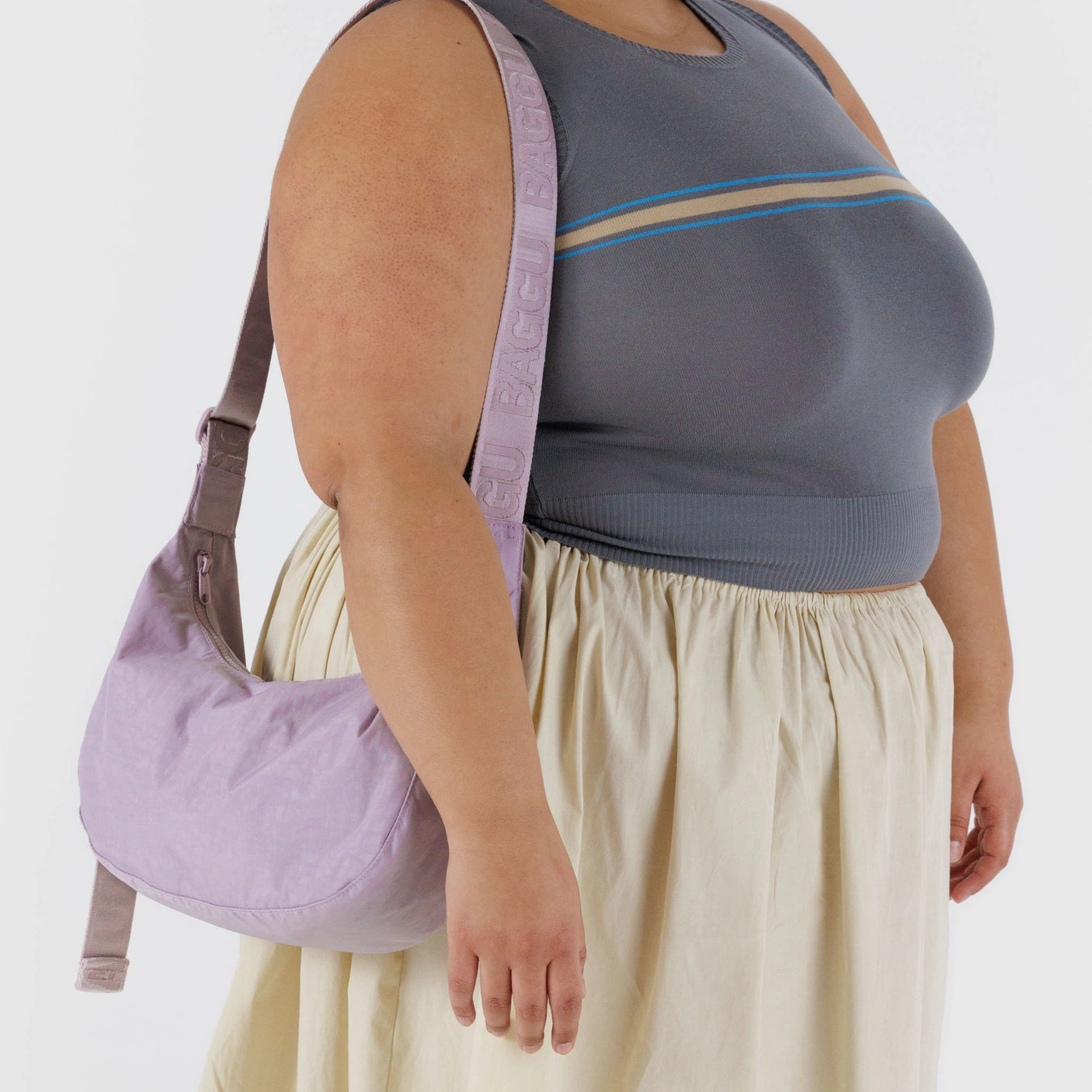 A person in a grey sleeveless top and cream skirt carries the Baggu Medium Nylon Crescent Bag in dusty pink over their shoulder, featuring an adjustable strap.