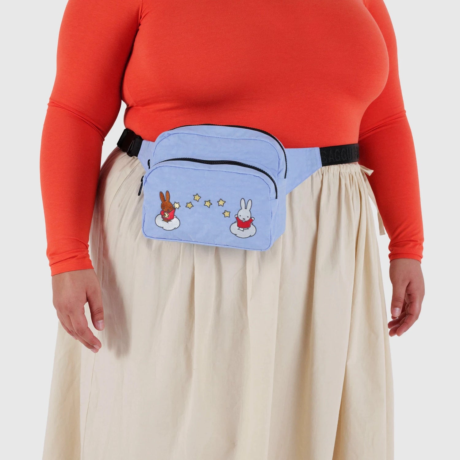 A person wearing a red-orange long sleeve top, cream skirt, and a blue Baggu Miffy x Baggu Fanny Pack made from recycled nylon, featuring cartoon bunnies and stars.