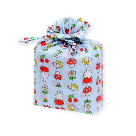 A rectangular gift wrapped in light blue cloth decorated with cartoon rabbits, flowers, mushrooms, and cherries, tied with a knot—evoking the playful design of the Baggu x Miffy Standard Baggu set of 3 bags by Baggu.