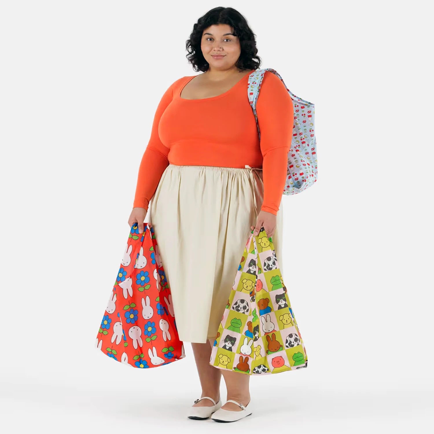 A person in an orange top and beige skirt carries the Baggu Standard Baggu x Miffy - Set of 3 Bags, with two colorful bags in hand and a third tote over their shoulder.