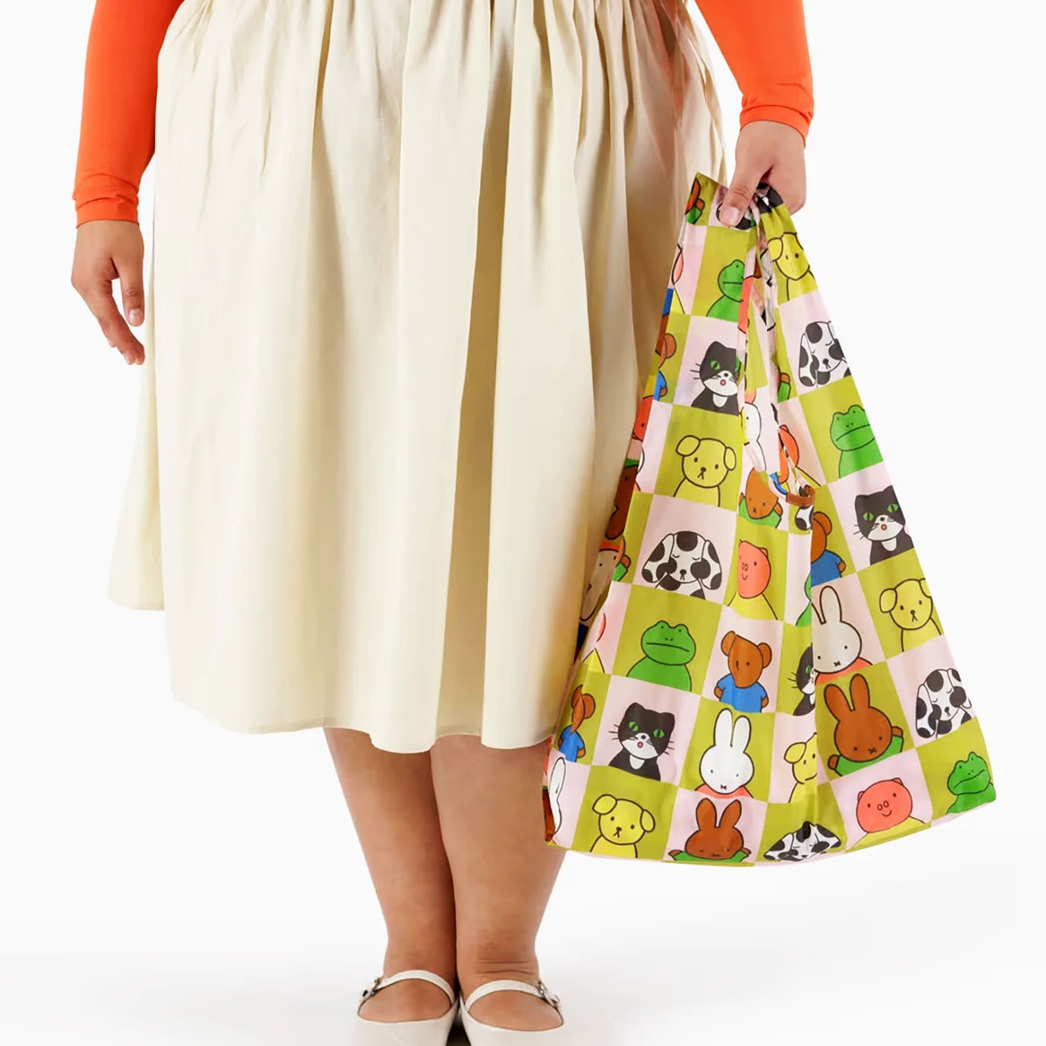 A person in a cream skirt and orange top holds a colorful Baggu x Miffy reusable bag featuring a grid of animal cartoon faces from the Standard Baggu x Miffy Set of 3 by Baggu.