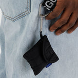 A Baggu Puffy Earbuds Case - Black is secured to a keyring loop on denim. A hand with dark skin is visible near the case.