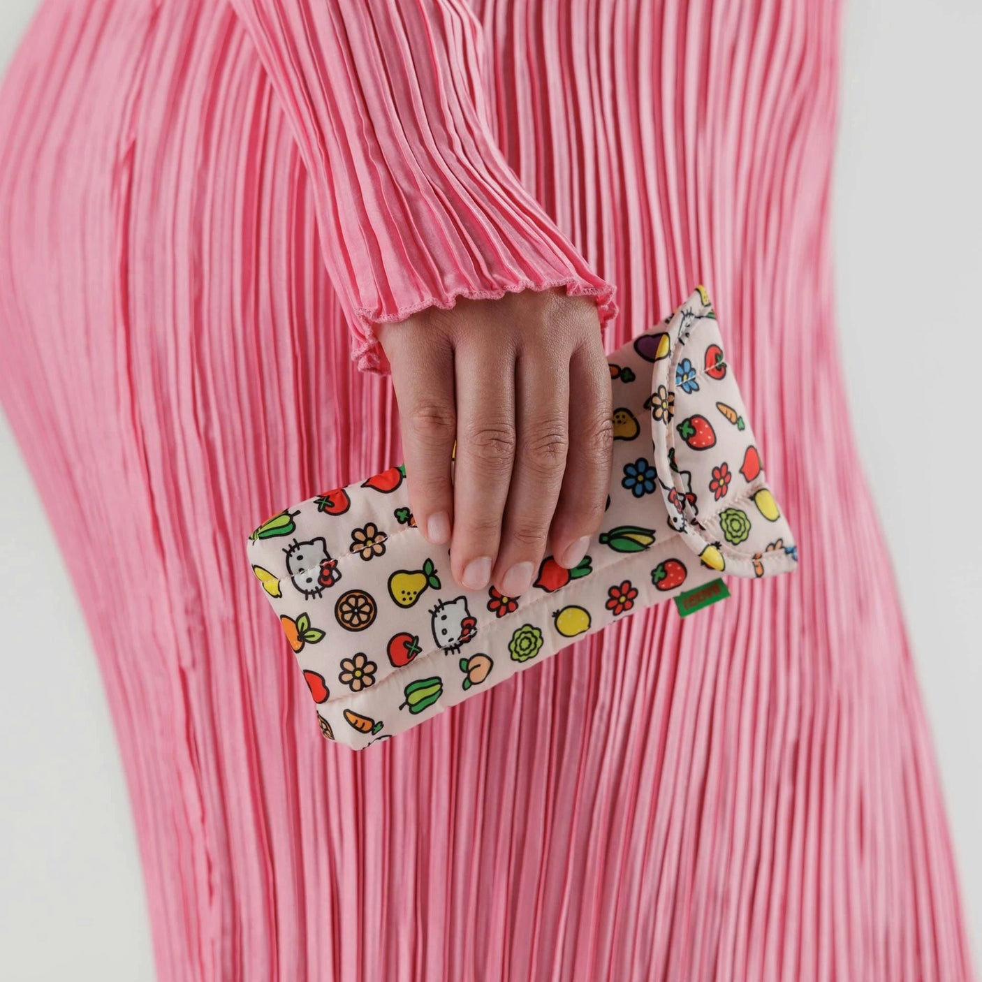 A person in a pink pleated dress holds a Baggu Glasses Sleeve featuring Hello Kitty Icons, adorned with colorful cartoon fruits and flowers, adding playful charm to the ensemble.