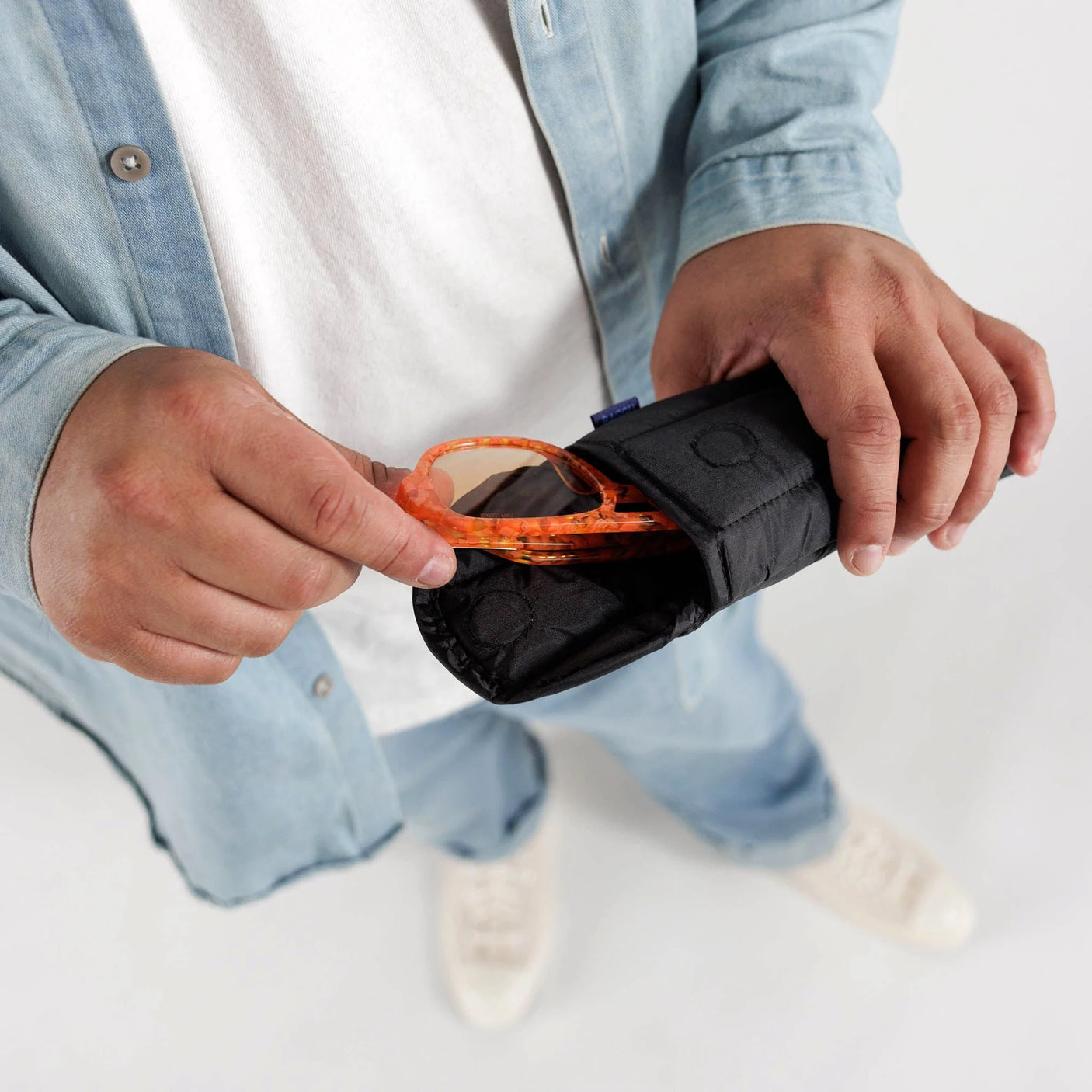 A person in a casual denim outfit holds a sleek Baggu Glasses Sleeve in black, stylishly placing orange sunglasses into the recycled nylon protective eyewear case.