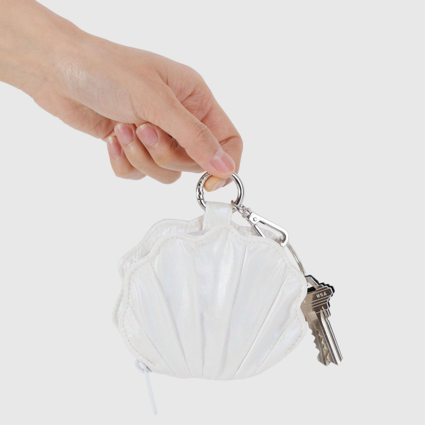 A hand holds a Baggu Shell Charm - Pearl Metallic keychain with two keys, an elegant white accessory, against a plain background.