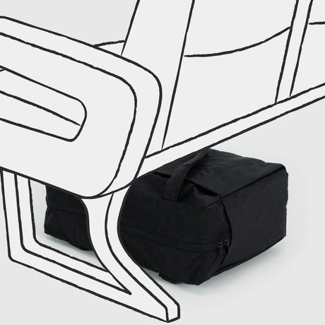 A Baggu Small Cloud Carry-on in black, crafted from recycled heavyweight nylon, is positioned under a stylized, partially drawn chair against a light gray background.