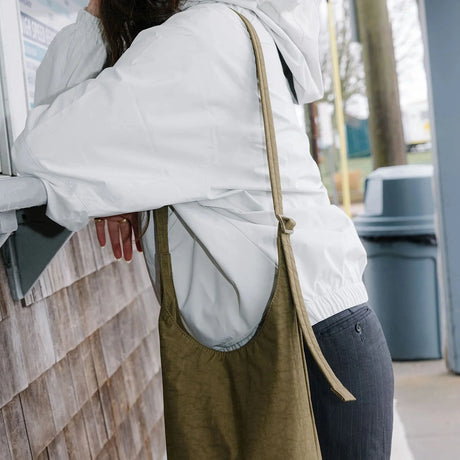 A person in a white jacket and black pants stands at a service window, carrying a Baggu Small Nylon Sling in the Seaweed color with an adjustable strap.