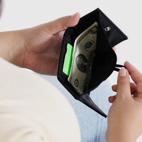 A person sits with a minimalist focus on their hands, holding an open Baggu Snap Wallet - Black from Baggu, revealing cash and a green card. The design harmoniously merges style and practicality while emphasizing functionality.