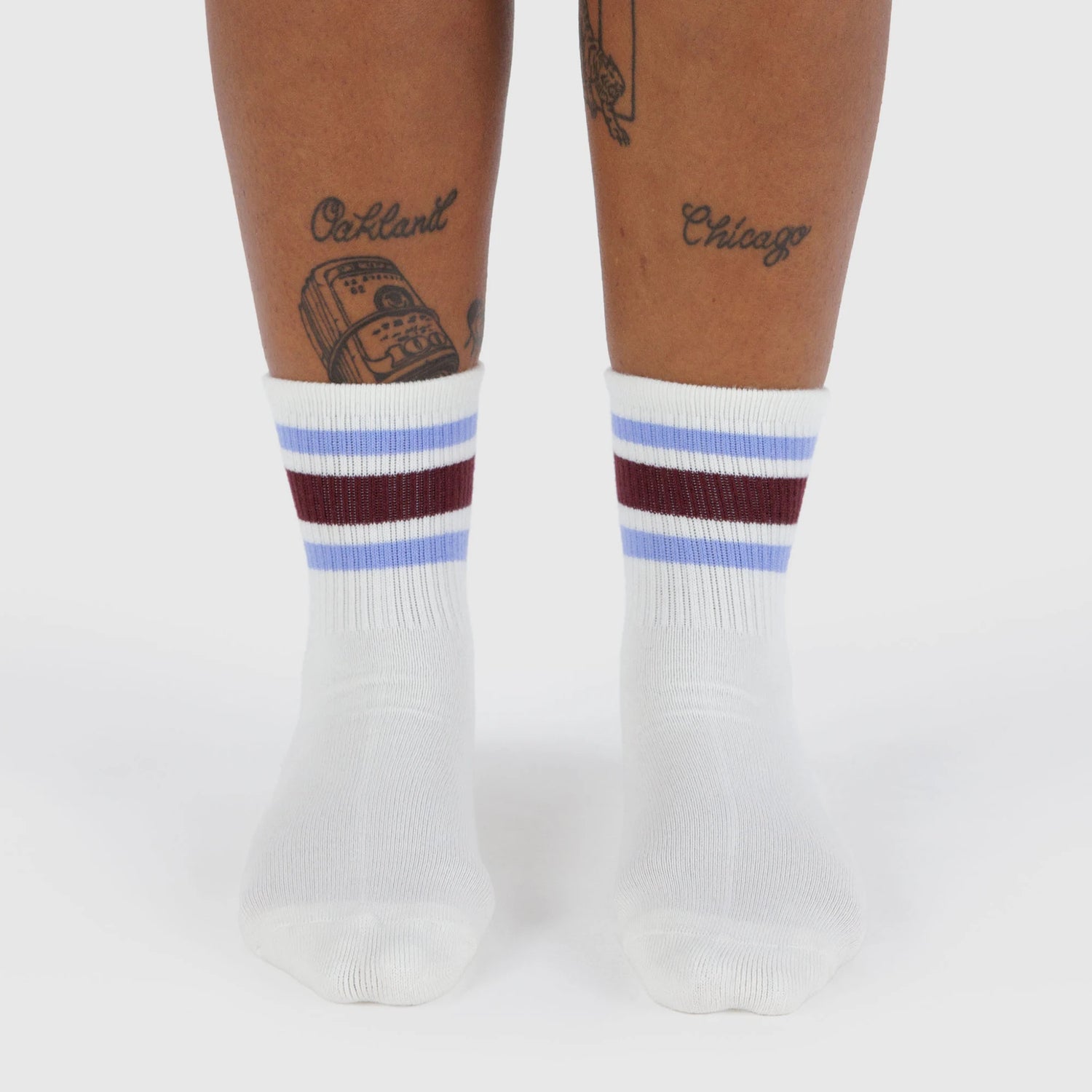 A person with leg tattoos wears Baggu Ribbed Sock - Ivory Stripe by Baggu—white crew socks with blue and burgundy stripes—standing on a white background.