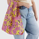 A person holding the Baggu Standard Reusable Bag - Rose, adorned with a vibrant pink rose pattern, and wearing a beige top and blue jeans.