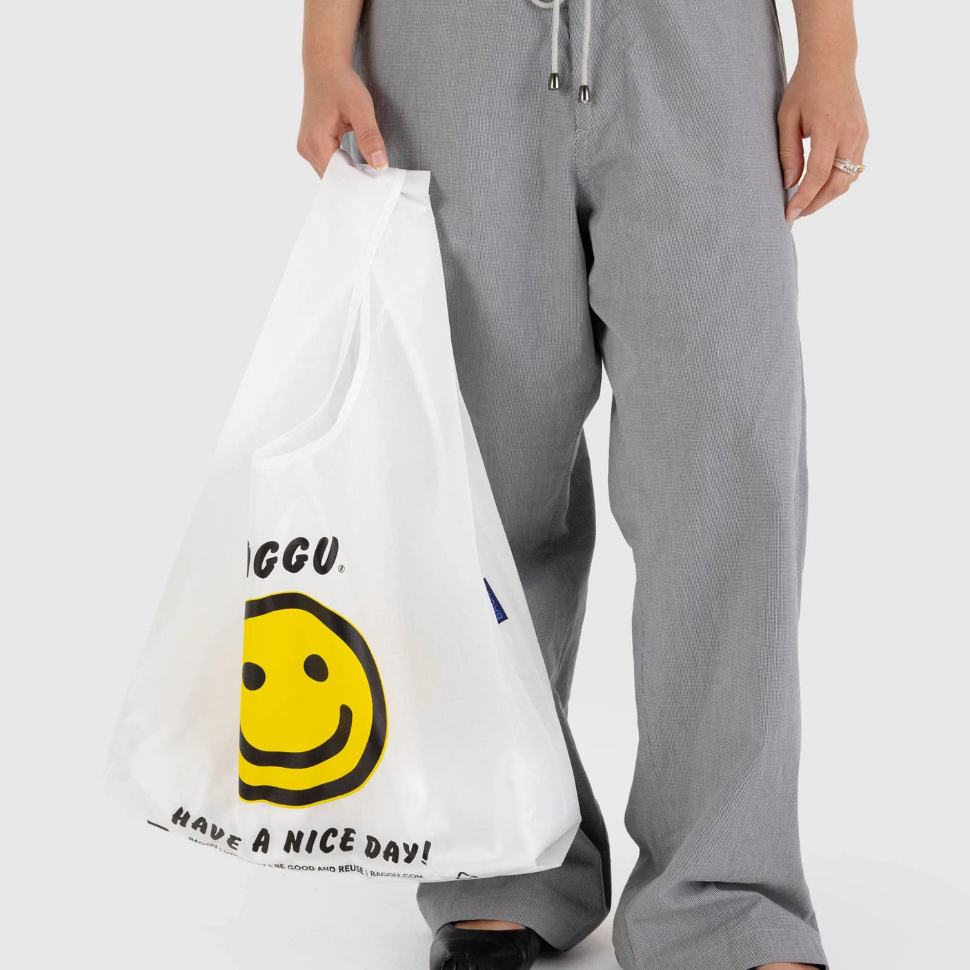 A person carrying the Baggu Standard Reusable Bag - Thank You Happy, crafted from recycled nylon, featuring a cheerful yellow smiley face and the text "HAVE A NICE DAY!" While gray pants peek through, this trendy accessory by Baggu is both sustainable and fashionable.