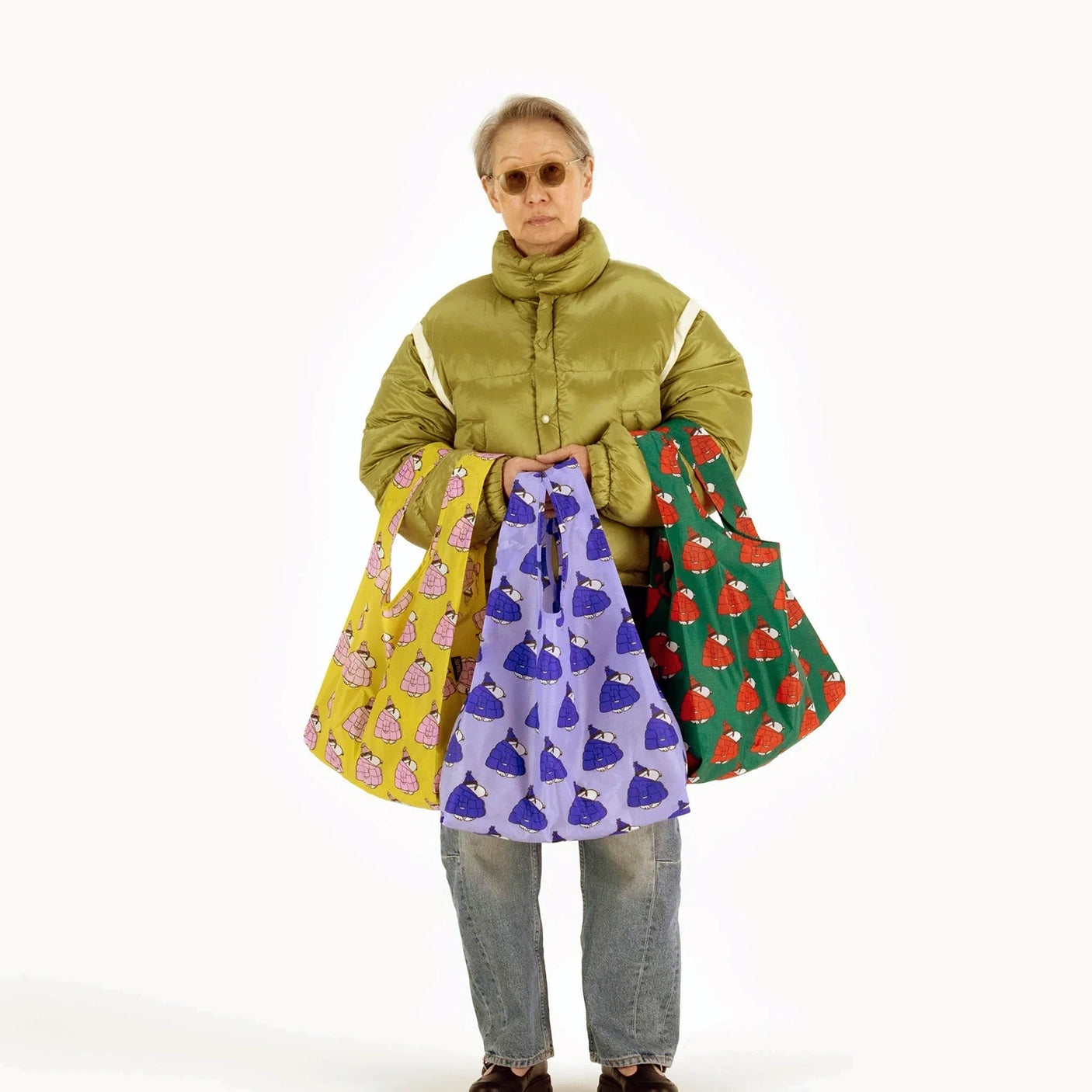 A person with sunglasses and a green jacket is holding three colorful, patterned bags from the Peanuts x Baggu Set of 3 Standard Bags, featuring Puffer Snoopy Prints. The backdrop is plain white.