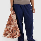 A person showcases sustainable style with the Baggu Standard Reusable Bag - Pointe Shoe pattern, paired with a beige top and blue pants, against a plain backdrop.