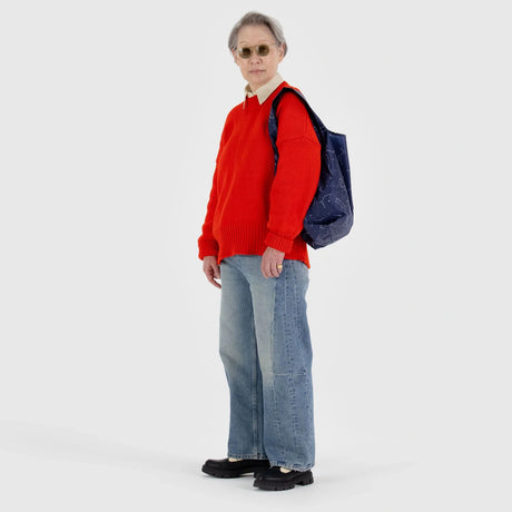 A person wearing a red sweater, blue jeans, and sunglasses carries a Baggu Standard Reusable Bag in the Constellation design, made from recycled nylon.