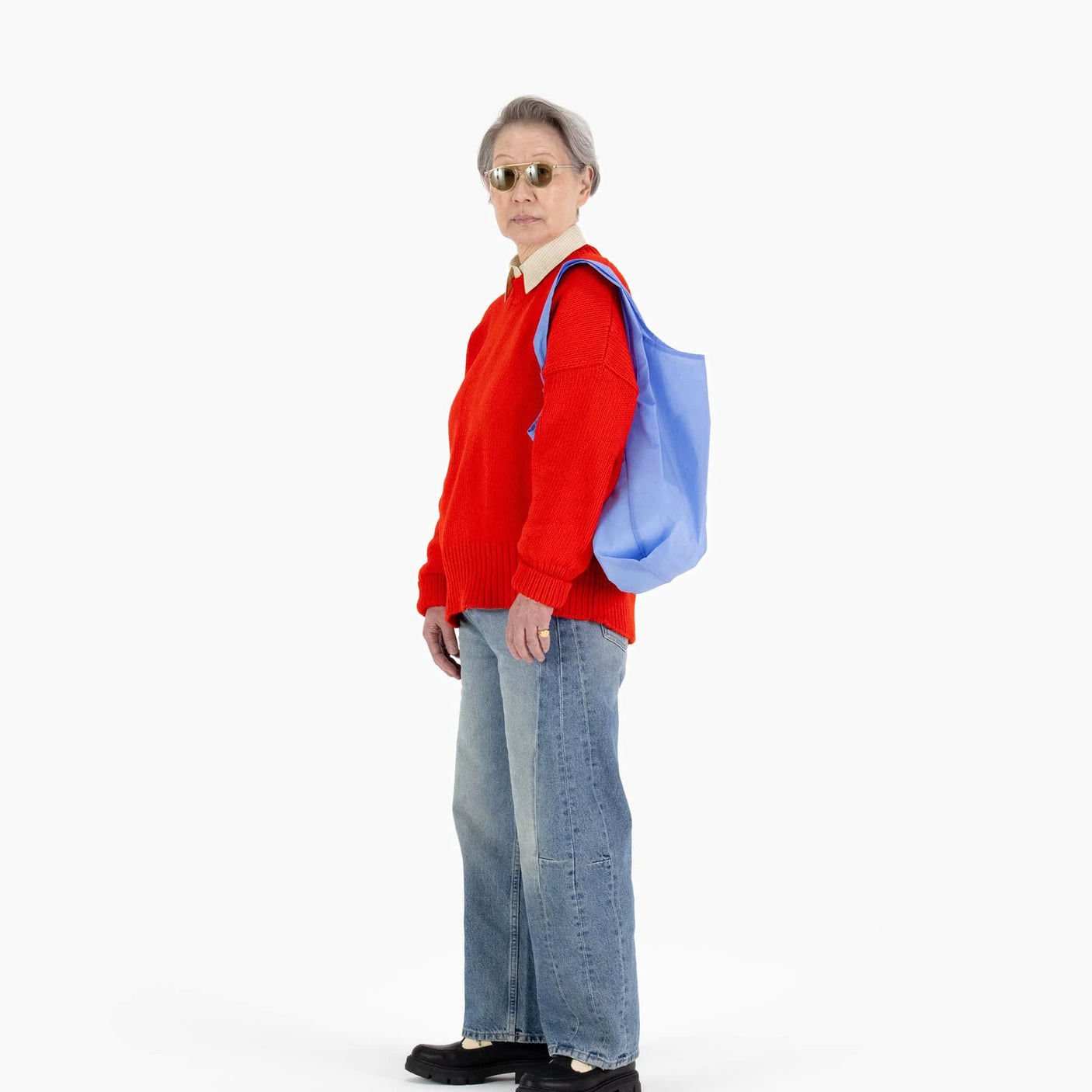 A person in sunglasses is dressed in a red sweater, jeans, and black shoes, while carrying a Baggu Standard Reusable Bag in Cornflower over their shoulder, against a plain white background.