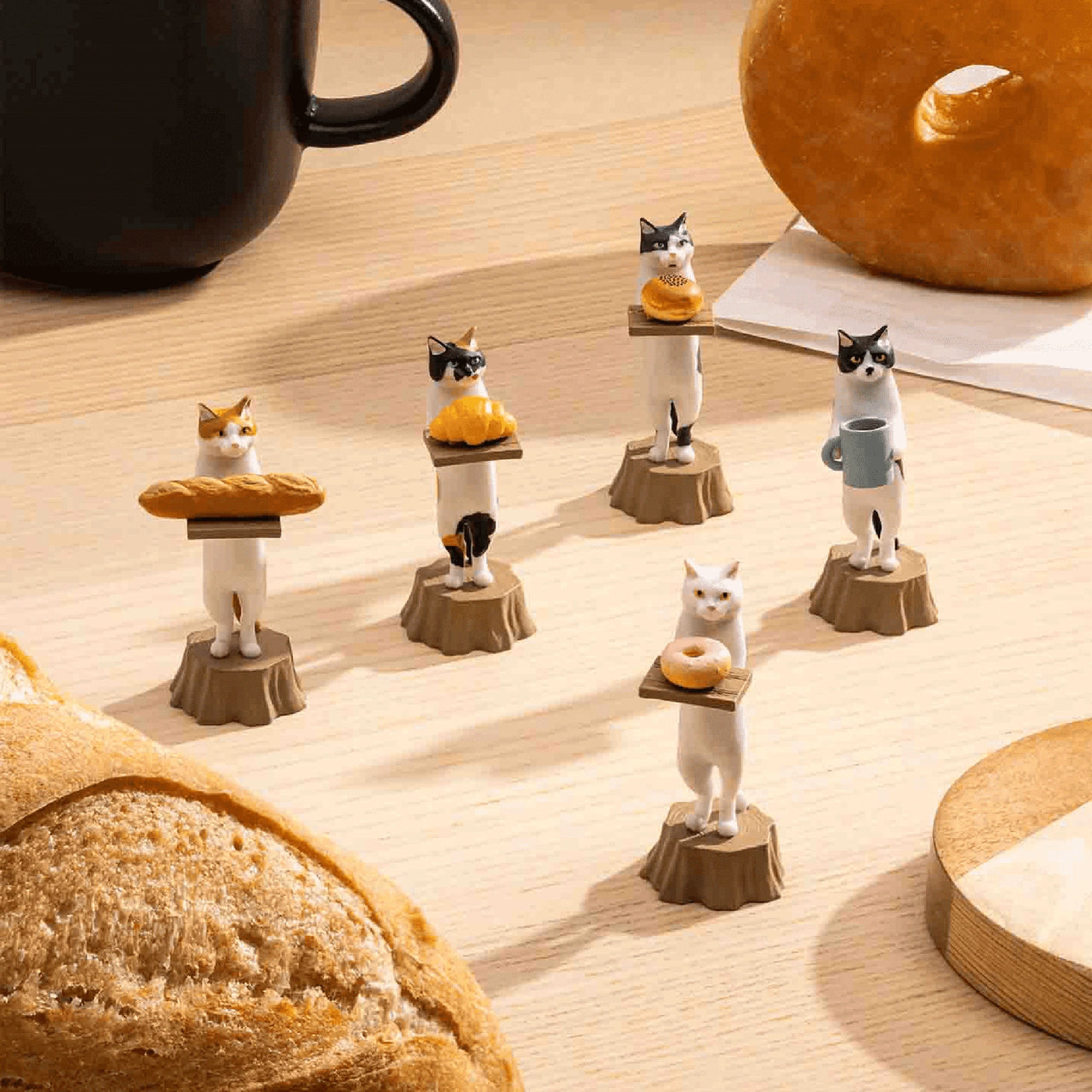 Five cat figurines from Kitan Club's Cat Bakery Waiter Blind Box stand on wooden bases, each holding a bakery treat: a bread loaf, croissant, donut, coffee, and a platter. A black mug complements the scene with additional bread on the wooden table.