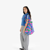 A person with long hair stands, dressed in a blue shirt and gray pants, carrying an eco-friendly Big Baggu Bag - Pink Apples & Bananas from the brand Baggu.