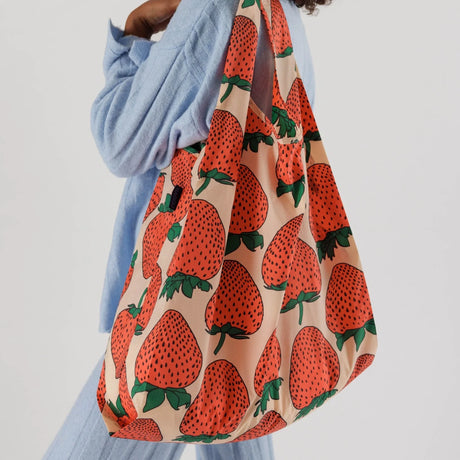 A person holding a Baggu Big Baggu Bag - Strawberry, an oversized beige tote adorned with a charming strawberry pattern and crafted from recycled nylon, pairs it perfectly with their light blue outfit.