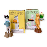 Two cat waiter figurines from the Kitan Club Cat Bakery Waiter Blind Box hold treats: one with a pastry and a blue mug, the other with a green dessert. The scene includes decorative packaging with warnings.