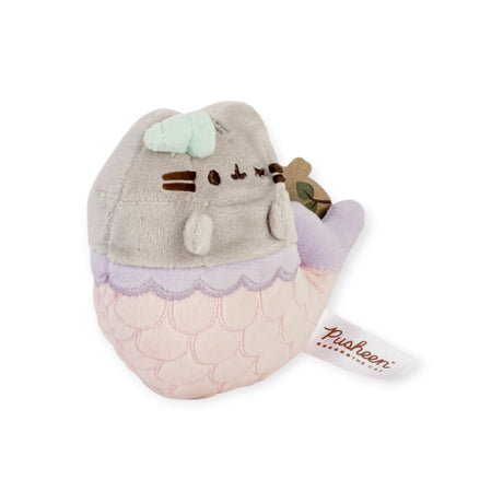 This 13cm Pusheen Unicorn & Mermaid Plush showcases a gray and pink cat with a mermaid tail, accented by a small green bow, holding a brown item. It's labeled on the side, making it an adorable collectible for fans of the Pusheen brand.