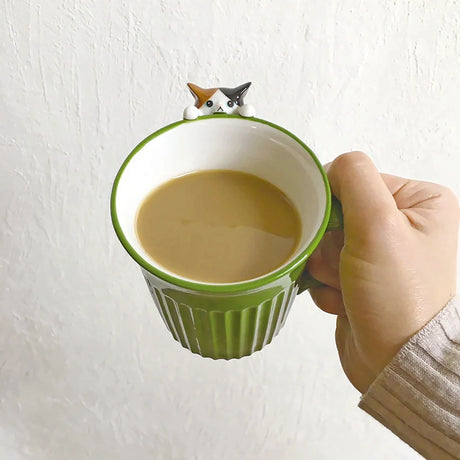 Hand holding green ceramic mug with white interior; calico cat figure sits on rim