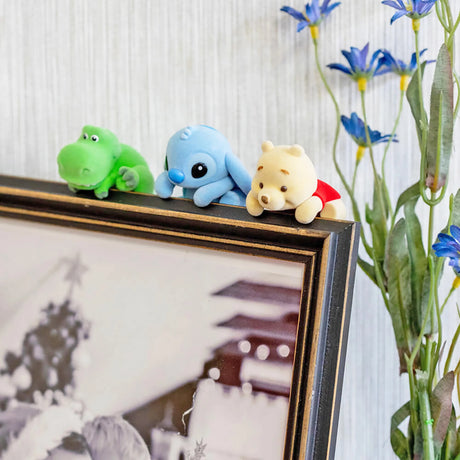 Three small colorful plush characters perched on the corner of a framed black-and-white photo with blue flowers nearby