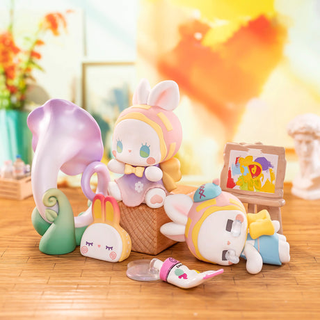 Two pastel bunny figurines from the Lucky Emma Fairy Babe Academy Blind Box pose in an art studio with a flower, easel, paint tube, and bunny pillow—ideal for magical school collectible fans.