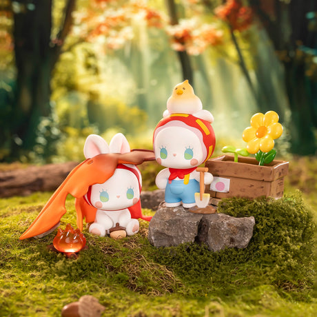 Two adorable figurines from the Lucky Emma Fairy Babe Academy Blind Box—one with bunny ears and an orange cape by a campfire, and one in a red hood with a shovel and bird—stand on moss near a flower box in a magical forest.