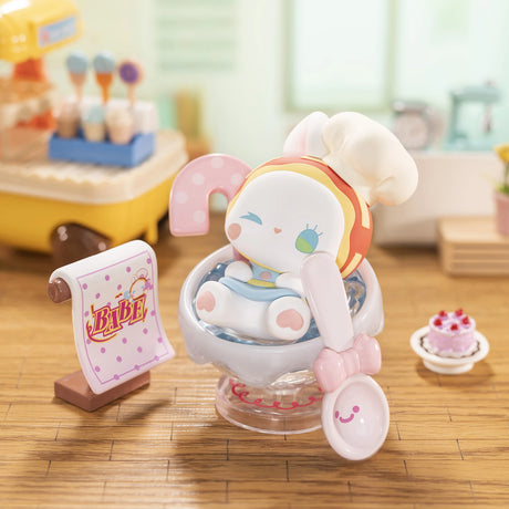 A cute toy bunny with a chef hat sits in a mixing bowl amid baking props on wood, cake, and recipe scroll—a whimsical addition for Lucky Emma fans and Emma Fairy Babe Academy Blind Box collectors.
