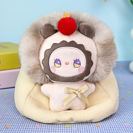 A plush toy with a round face, faux fur hood, and red pom-pom sits on a cream cushion with a yellow bow—the Emma Fuzzy Family Bag Charm - Blind Box by Lucky Emma, an adorable animal bag charm perfect for collectors.