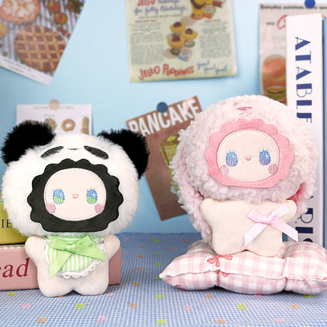 Two pastel plush dolls with round faces are shown on a dotted surface; one wears a panda hat like the Lucky Emma Fuzzy Family Bag Charm - Blind Box, while the other has a fluffy pink hood and sits on a checkered pillow—great as animal bag charms.