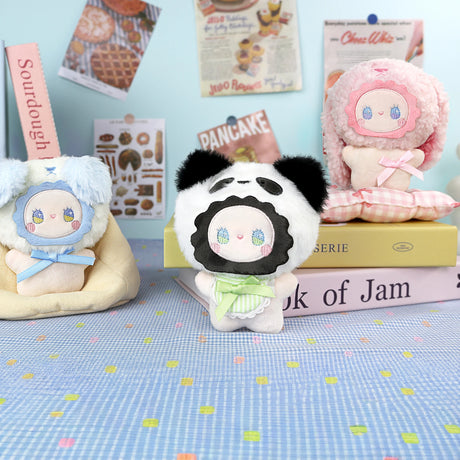 Three Emma Fuzzy Family plush dolls by Lucky Emma, each with animal-themed hoods and pastel colors, are displayed on a table with books as food-themed posters decorate the backdrop; each doubles as a plush keychain for your bag.