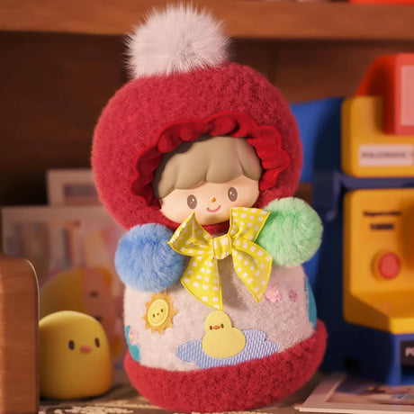 Plush keychain featuring a character in a red hood with pom-poms and a yellow bow, surrounded by toys on a desk