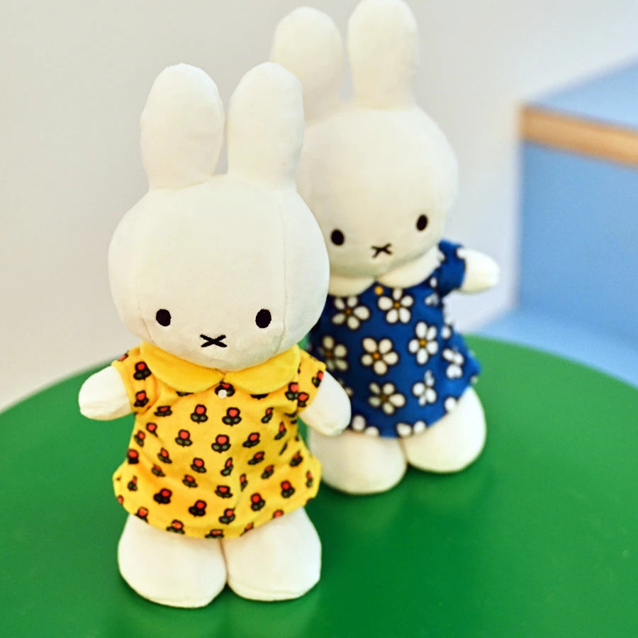 Two Miffy plush toys with white fur stand on a green surface; one wears an exclusive yellow floral dress from the "Miffy Flower Dress Plush 23cm - Limited Edition" collection, and the other is dressed in a blue daisy outfit.