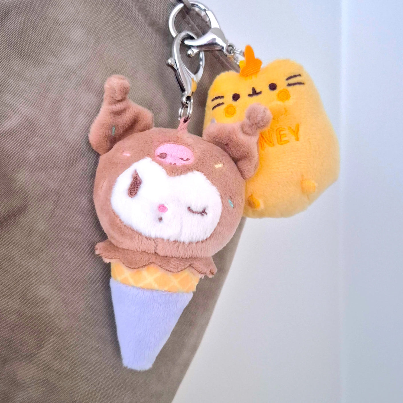 Two charming plush keychains featuring Hello Kitty & Friends: one resembling an ice cream cone with a snoozing bear face and the other shaped like a yellow cat adorned with "HONEY." They hang from a metal clip on the fabric surface, reminiscent of delightful Hello Kitty plush toys.