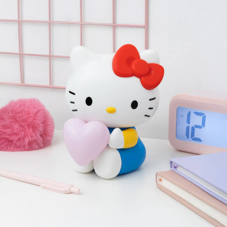 A Hello Kitty Night Light from the Hello Kitty brand, featuring a figure holding a glowing pink heart, sits on a desk beside a pink fluffy object, a pen, and a clock displaying the time 12:00.