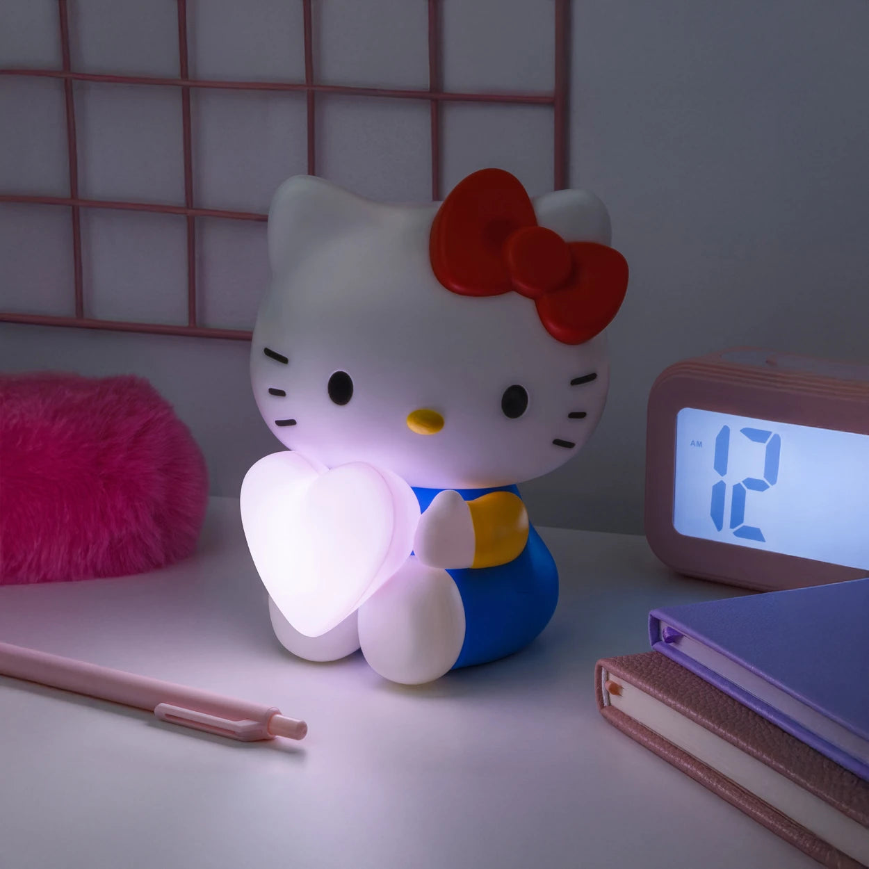 A Hello Kitty Night Light (16cm) from the Hello Kitty brand, featuring a glowing pink heart, sits on the desk, illuminating the space alongside a digital clock, a pink pen, and a book.