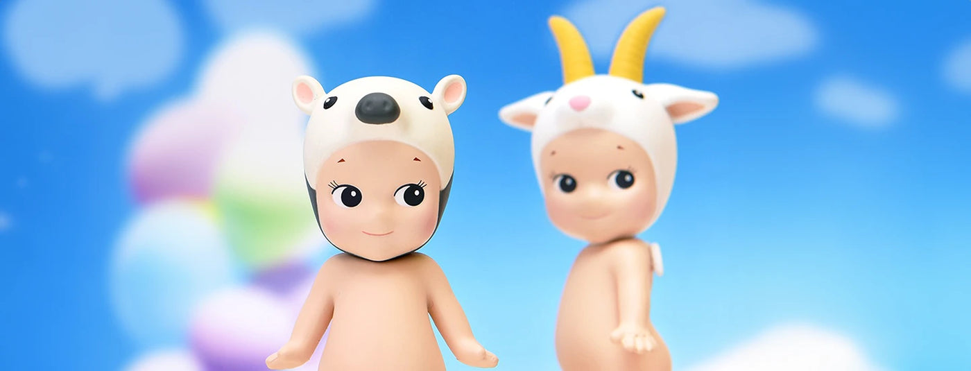 Two figurines with animal-themed hats, one with a bear design and the other with a goat design, standing against a colorful cloud-filled sky background.