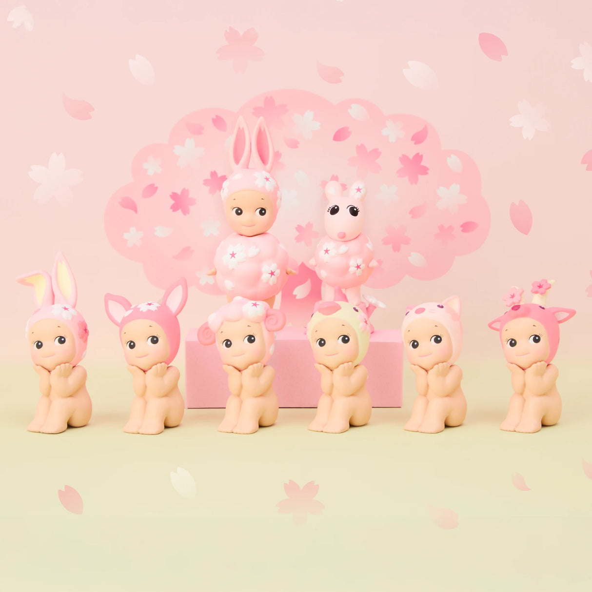 Part of the limited edition Sonny Angel Cherry Blossom Series, seven baby figurines donning cute animal hats are displayed. Two extras sit on a pink box: one dressed as a rabbit, the other with a fluffy collar, all set against cherry blossom motifs—ideal for collectors.