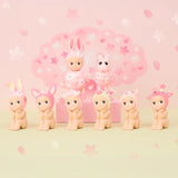 Part of the limited edition Sonny Angel Cherry Blossom Series, seven baby figurines donning cute animal hats are displayed. Two extras sit on a pink box: one dressed as a rabbit, the other with a fluffy collar, all set against cherry blossom motifs—ideal for collectors.