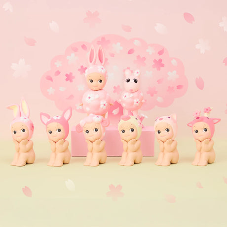 Part of the limited edition Sonny Angel Cherry Blossom Series, seven baby figurines donning cute animal hats are displayed. Two extras sit on a pink box: one dressed as a rabbit, the other with a fluffy collar, all set against cherry blossom motifs—ideal for collectors.